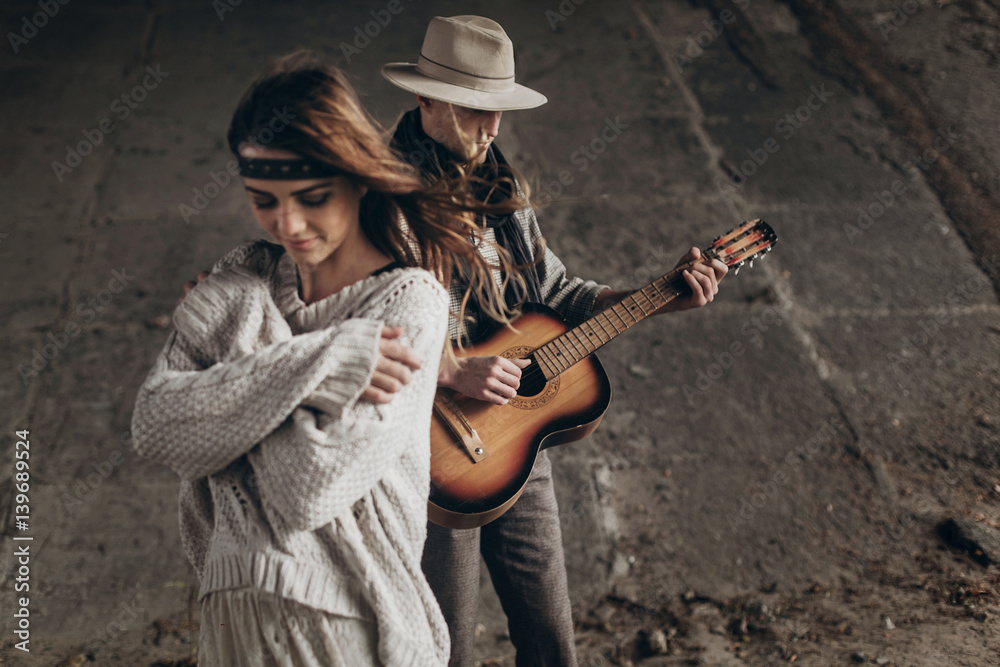 stylish hipster couple posing. man in hat playing guitar for his boho woman in knitted sweater. atmospheric sensual moment. rustic fashionable look.