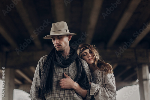 stylish hipster couple dreaming under abandoned bridge. boho gypsy woman gently hugging arm of confident man in hat. atmospheric sensual moment. rustic fashionable look.