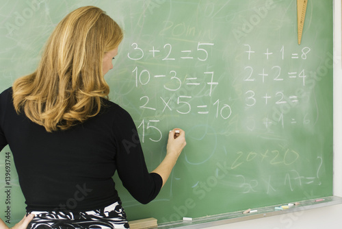 Elementary teacher writing arithmetic on blackboard, rear view