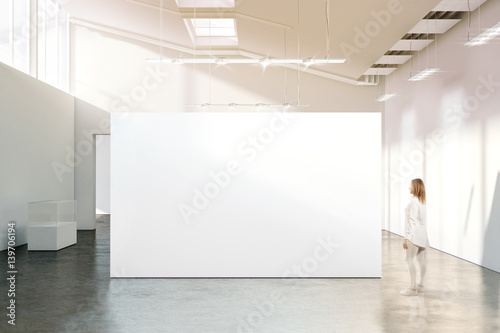Woman walking near blank white wall mockup in modern gallery. Girl admires a clear big stand mock up in museum with contemporary art exhibitions. Large hall interior, banner exposition show