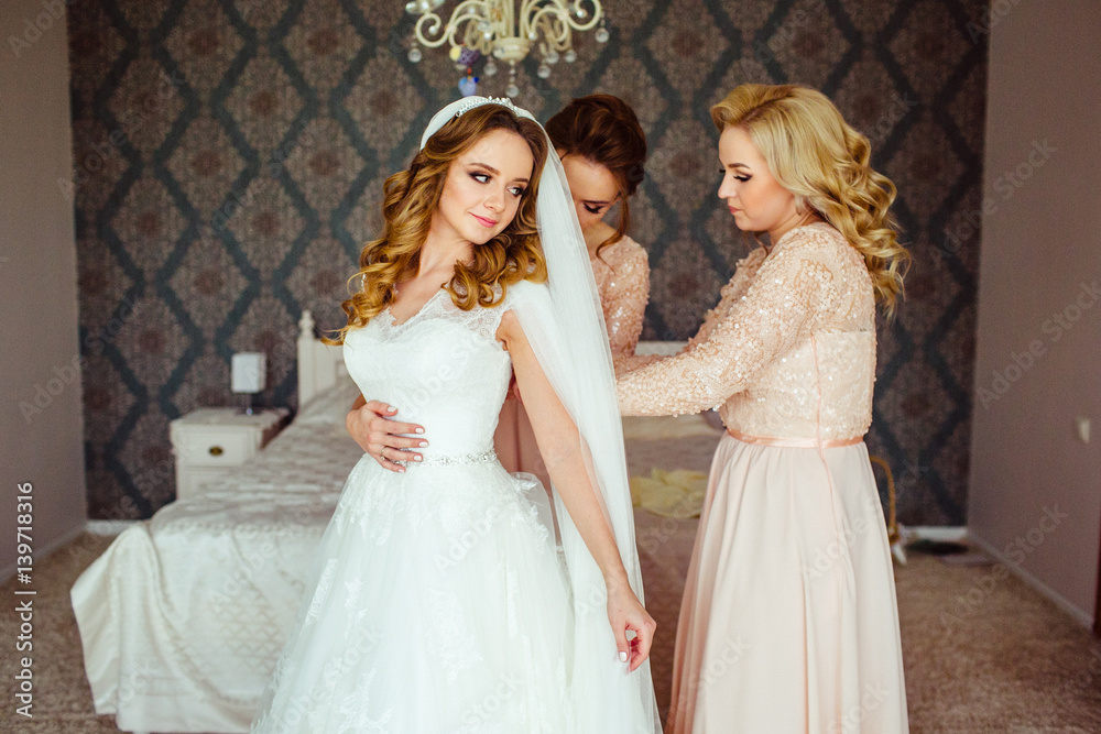 Bridesmaids in pink dresses help bride to put on wedding dress