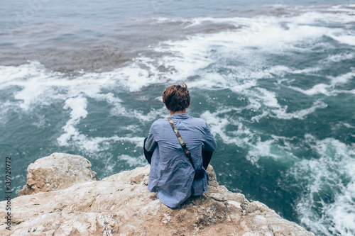 A young adventurous man sitting on the top of the cliff overlooking the pacific ocean