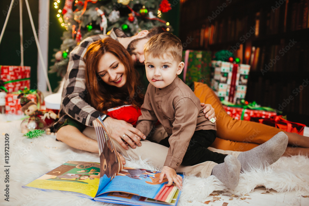 Funny little child reads colorful book with his parents before Christmas tree