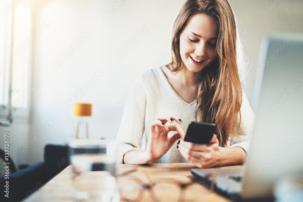 Happy smiling young girl send an sms message to her best friend while working at modern office, cheerful woman using smartphone at home interior, joyful hipster girl chatting via cellphone Happy smiling young girl send an sms message to her best friend while working at modern office, cheerful woman using smartphone at home interior, joyful hipster girl chatting via cellphone