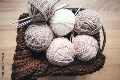 Beige yarn, knitting needles and a brown scarf are in the basket. Wooden background. Hobbies 