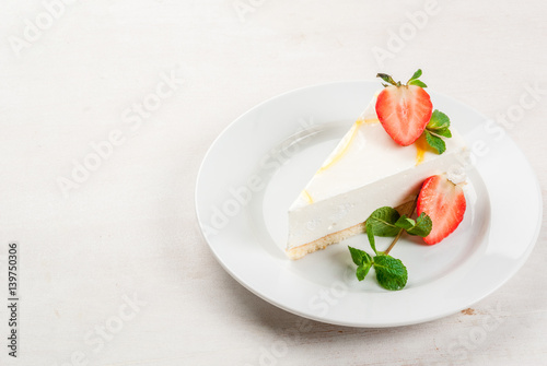 Light dessert: classic cheesecake, decorated with mint and strawberries. On a white plate, on a white wooden table. 
