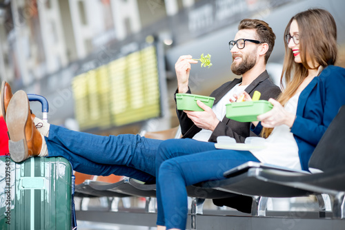 Elegant business couple eating with lunch boxes sitting at the waiting hall in the airport. Having a snack during business trip