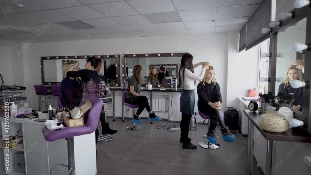 Professional hair stylist and makeup artist in their large studio preparing models before going on the podium for the show. Beautiful girl admires itself in a mirror and then looks in the smartphone. Professional hair stylist and makeup artist in their large studio preparing models before going on the podium for the show. Beautiful girl admires itself in a mirror and then looks in the smartphone.