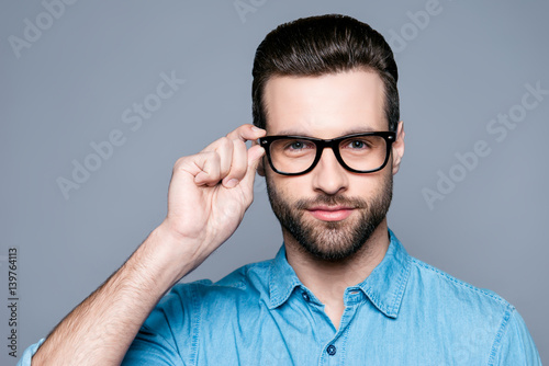 A portrait of young handsome man in jeans shirt  isolated on gray background touching his glasses