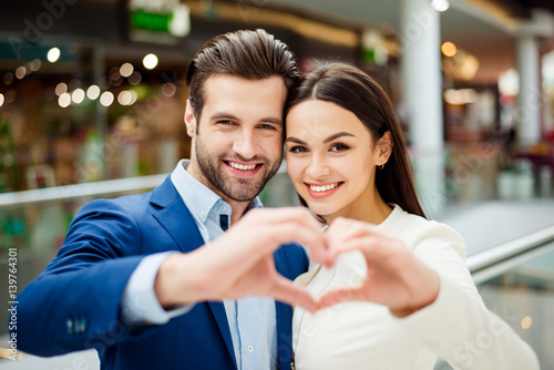 Happy lovely young woman with her handsome boyfriend  in suit together holding and showing hands in heart sign. Sweet couple falling in love, walk in modern mall and having fun at date.