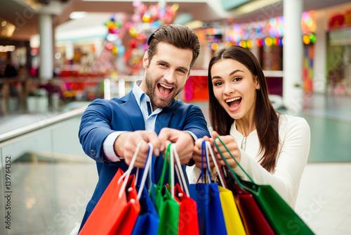 It's shopping and fun time. Portrait of cheerful successful happy young lovely couple holding colored shopping bags and laughing in mall. Concept of consumerism, sale, rich life and people