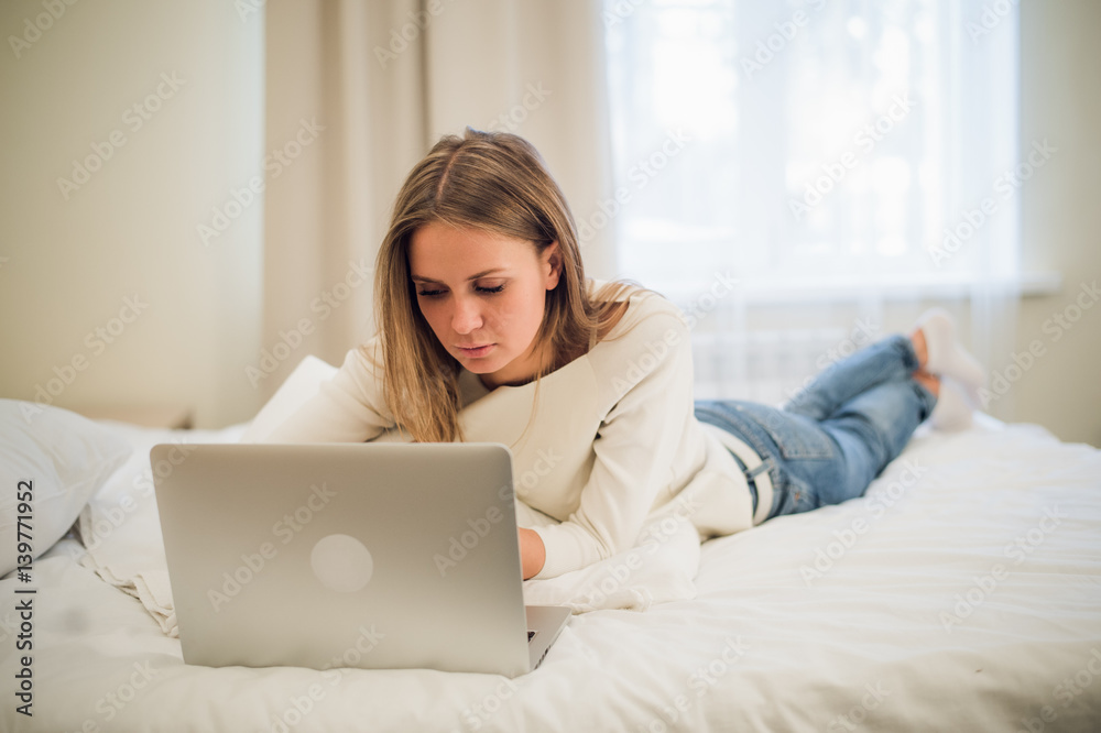 A cute young woman smiling excitedly at her laptop screen. She is relaxing in bed. A cute young woman smiling excitedly at her laptop screen. She is relaxing in bed.