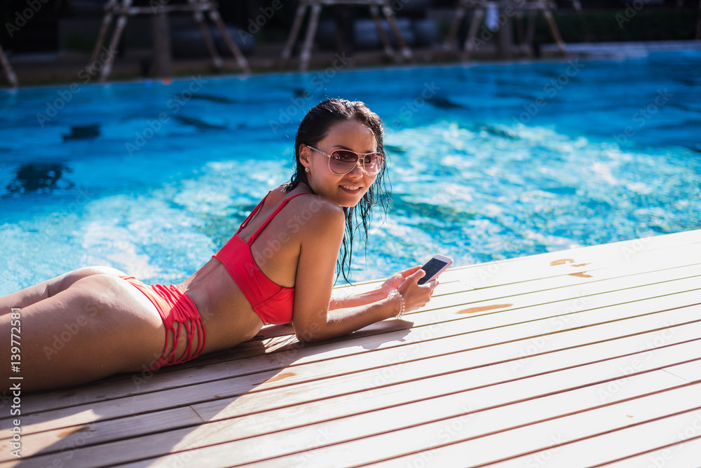 Beautiful woman leaning on poolside and typing a text message on cellphone