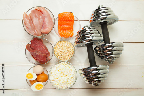 Protein diet, fish, cheese, eggs, meat, chicken and dumbbells on a white wooden background