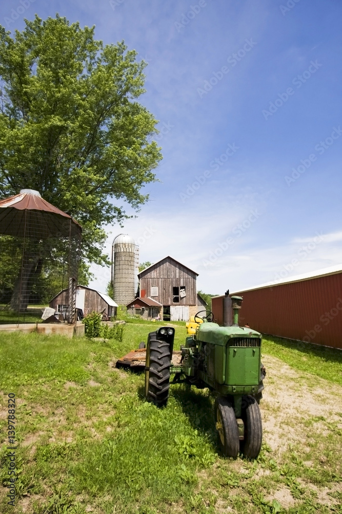 Obraz premium Agricultural and rural life background. Summer countryside landscape with old farm buildings and tractor at old farm in Wisconsin, Midwest USA. Beautiful sunny day with cloudy blue sky. Vertical shot.