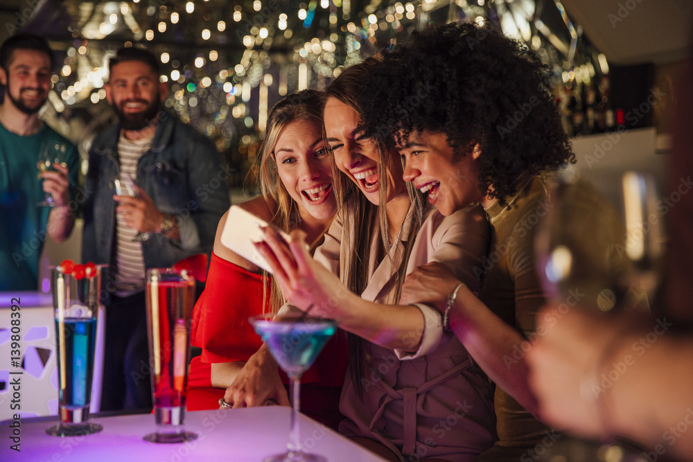Three women are taking selfies together on a smartphone while enjoying drinks in a night club. - obrazy, fototapety, plakaty 
