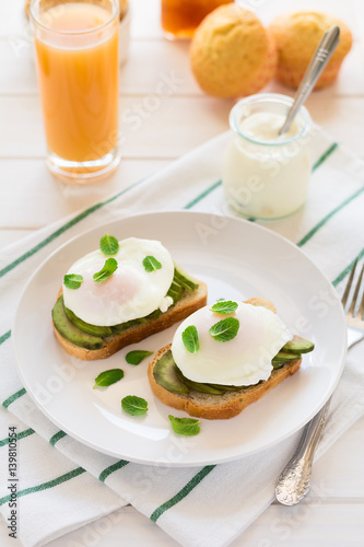 Vegetarian breakfast: avocado toast with poached eggs, orange juice, yogurt and jam on white wooden background. Selective focus