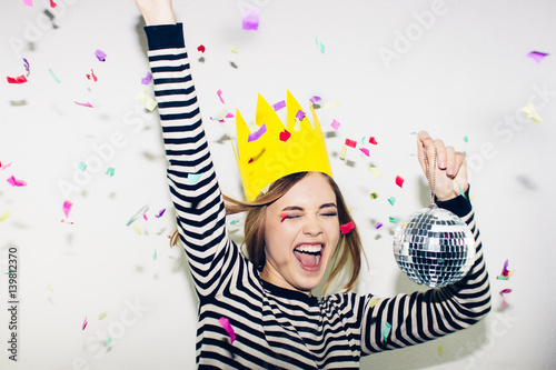 Birthday party, new year carnival. Young smiling woman on white background celebrating brightful event, wears stripped dress and yellow crown. Sparkling confetti, having fun, dancing, laugh, smile. photo