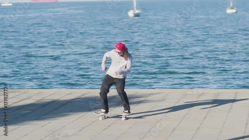 CLOSE UP SLOW MOTION: Young pro skateboarder skateboarding and jumping ollie flip trick on promenade along the coast on sunny summer day with beautiful green cape and the ocean in the background photo