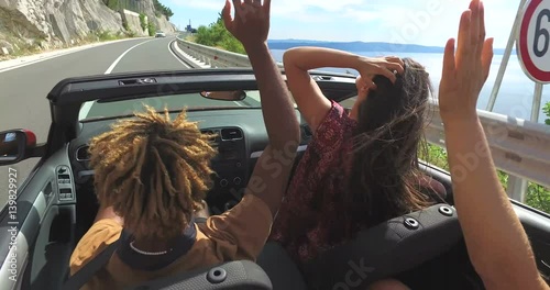 Friends dancing and waving arms driving on road along coast in red convertible