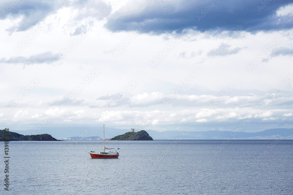 Immagine panoramica di una barchetta rossa in mezzo al mare su sfondo costa e cielo a tratti bianco e a tratti blu