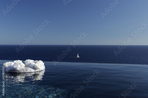 Foto di una piscina che si sovrappone al mare sullo sfondo. Nel primo piano c'è un ornamento floreale bianco e sullo sfondo un barca a vela bianca.