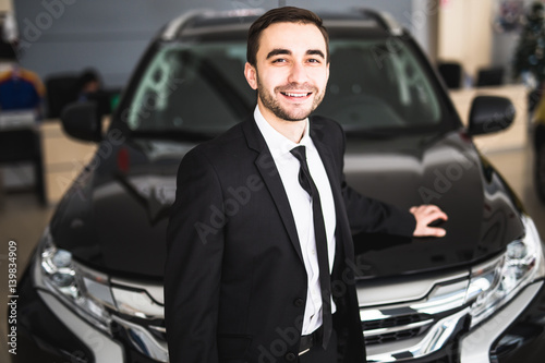 Handsome young classic car salesman standing at the dealership in front of new car