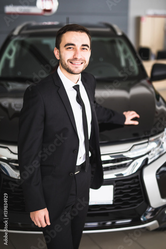 Handsome young classic car salesman standing at the dealership in front of new car photo
