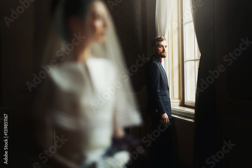 elegant stylish handsome groom looking at gorgeous bride, standing at window light. unusual luxury wedding couple in retro style