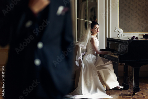 elegant gorgeous bride playing the fortepiano in rich old room and looking at stylish groom. man holding hand on suit. unusual luxury wedding in retro style photo