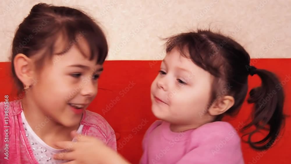 Kids whispering, two little girls indulge in playing on the orange couch. Two sisters hug. Kids whispering, two little girls indulge in playing on the orange couch. Two sisters hug.