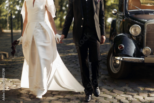 luxury elegant wedding couple walking and holding hands close up at stylish black car in light in park. gorgeous bride and handsome groom in retro style © sonyachny