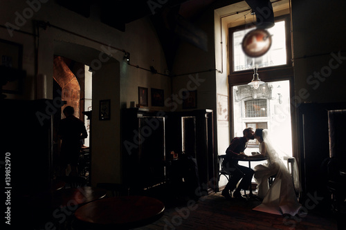 luxury wedding couple holding hands and kissing in cafe in morning near window in soft light. elegant gorgeous bride and stylish groom in retro style. romantic moment