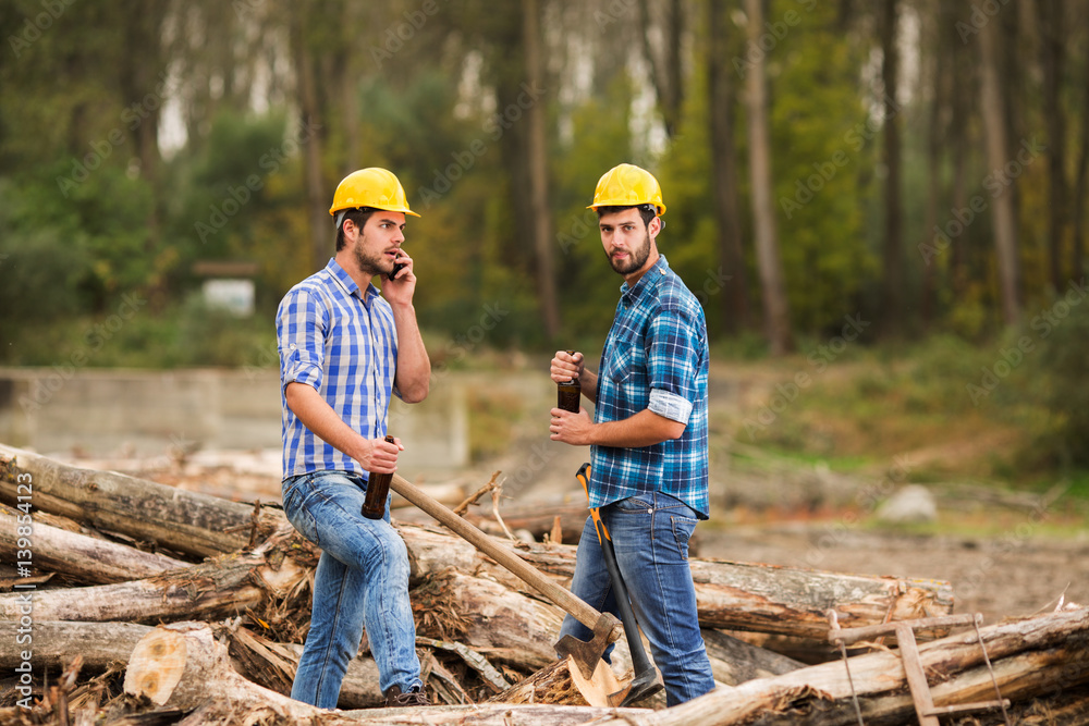 Two guys, with yellow helmets on their heads, rest in the forest after work and drink beer Two guys, with yellow helmets on their heads, rest in the forest after work and drink beer