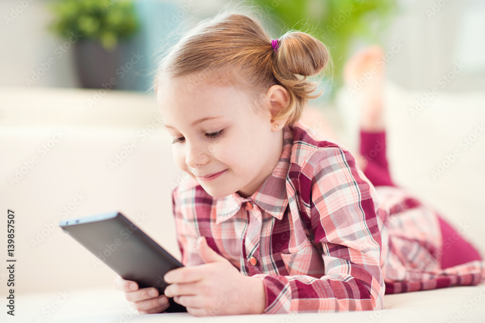 Pretty little child girl using a digital tablet, looking and smiling while lying on the whitw couch Pretty little child girl using a digital tablet, looking and smiling while lying on the whitw couch