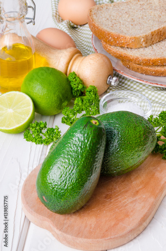 Fresh, green avocado, lime, parsley, sea salt, pepper, bread and eggs for sandwich on a white wooden background