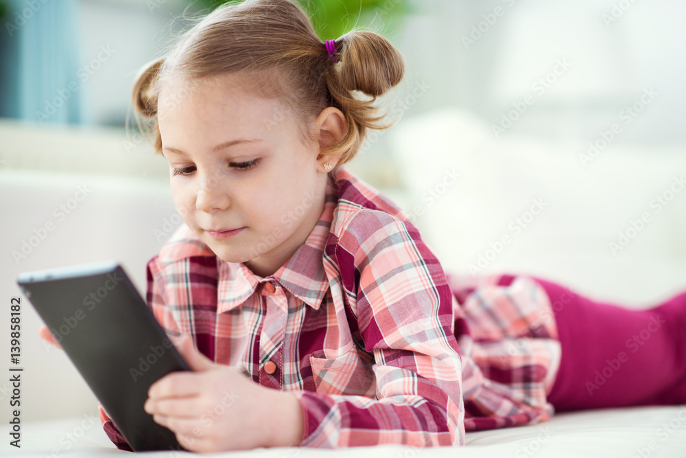 Pretty little child girl using a digital tablet, looking and smiling while lying on the whitw couch Pretty little child girl using a digital tablet, looking and smiling while lying on the whitw couch
