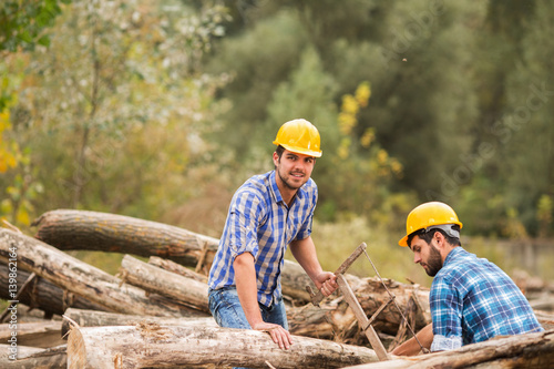 Two guys, with yellow helments on their heads, cut wood with a saw in the forest
