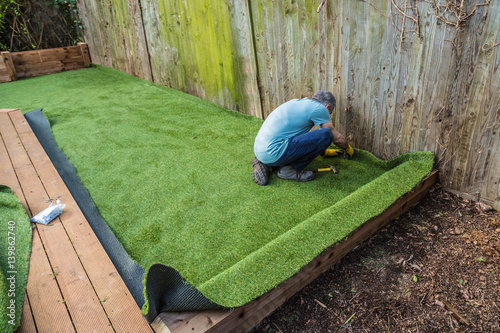 Artificial grass being installed, added next to wooden decking. It has been cut to size and rolled out and laid and is being nailed down. the installer has a hammer and nails.