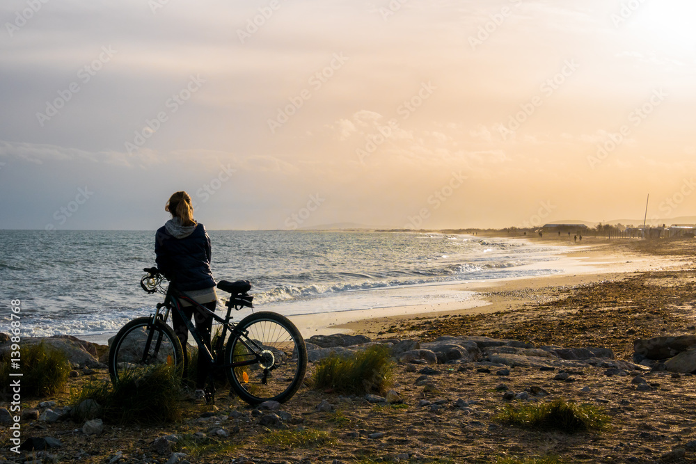 Fototapeta premium une femme appuyée sur son vélo, regarde un coucher de soleil sur une plage 