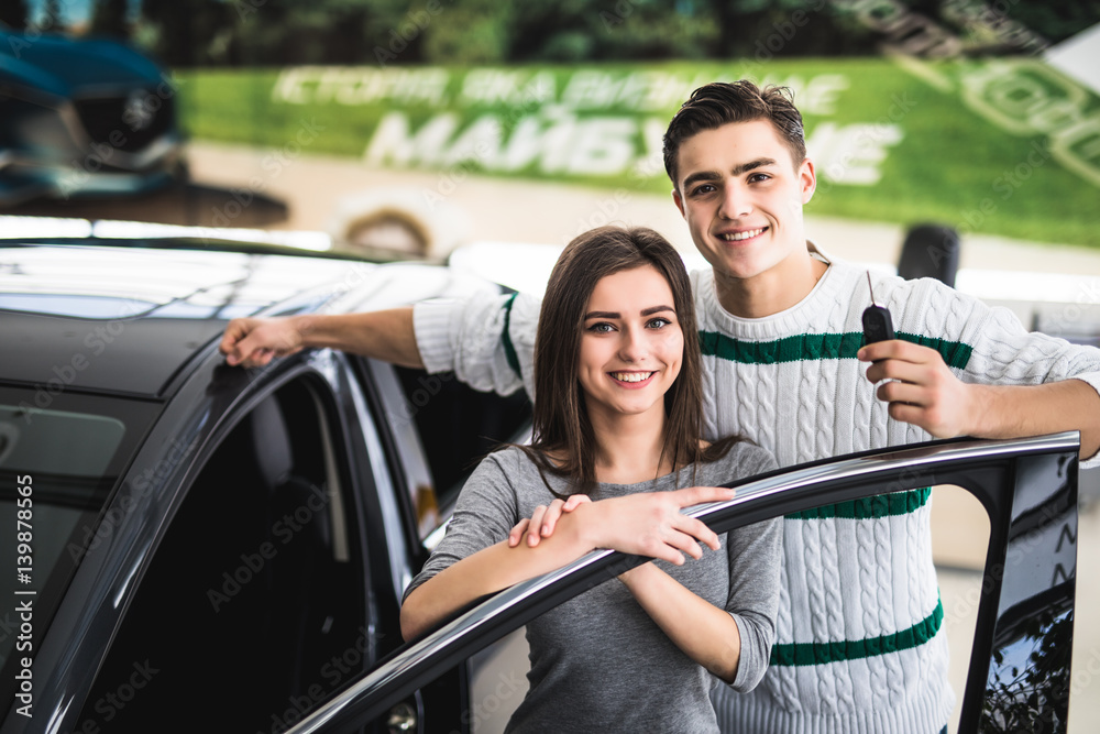 Beautiful young couple is smiling and looking at camera while leaning on their new car in a motor show. Man is holding car keys