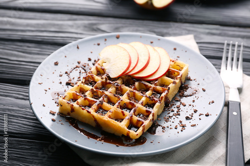 Delicious waffle with apple slices, syrup and chocolate shavings on plate