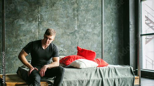 Young sexy men bodybuilder athlete,studio portrait loft on background of stylized wall,guy model black Tshirt and trousers Sits on bed with pillows and rug on brutal male bed made of metal and pallets
