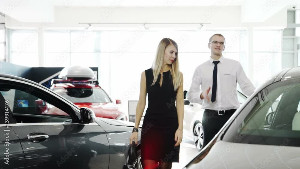 female customer comes to car, seller consultant tells about automobile. redhead tall male in white shirt, trousers, tie, glasses explains woman advantages, disadvantages of product in showroom centre female customer comes to car, seller consultant tells about automobile. redhead tall male in white shirt, trousers, tie, glasses explains woman advantages, disadvantages of product in showroom centre
