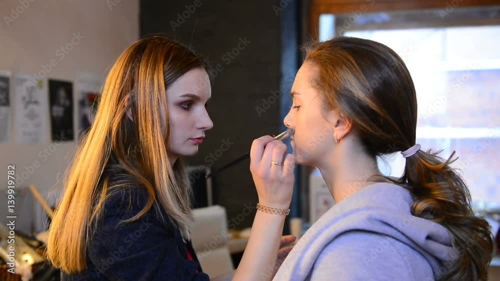At the beauty salon female makeup brush does makeup on the model face. Master with long red hair straight with the help of medium sized brush to apply foundation cream. She spends fluffy instrument on At the beauty salon female makeup brush does makeup on the model face. Master with long red hair straight with the help of medium sized brush to apply foundation cream. She spends fluffy instrument on