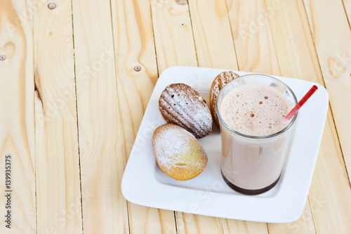 Chocolate smoothie, cocoa milkshake in the glass.French Madeleine cookies powdered sugar on the white plate. Light-colored natural wooden background. Copy space