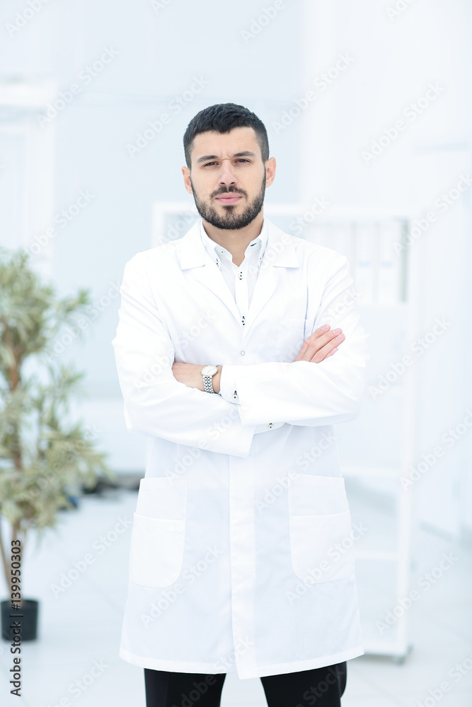 Smiling doctor posing with arms crossed in the office, he is wearing a medical staff on the background