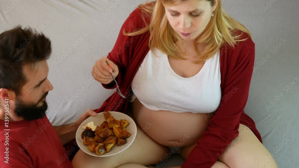 Time-lapse of pregnant loving couple eating potato wedges on the bed at home.