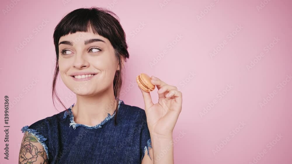 Young fresh and beautiful brunette girl isolated on side on pale pink background smiles, thinks about something, looks on sides and holds small macaron cookie in her hand, wearing denim dress