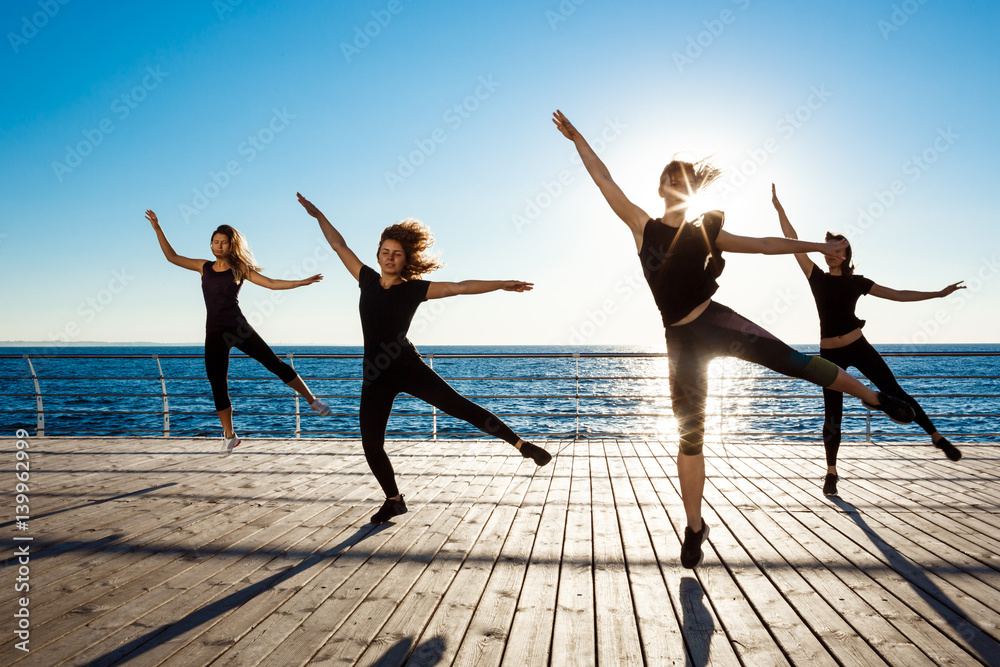 Fototapeta premium Silhouettes of sportive girls dancing near sea at sunrise.